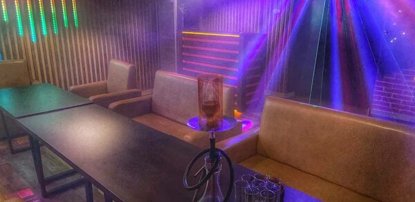 Forest(Лес). Тёмная комната с длинными столами, коричневыми диванами и креслами. На столе кальян и стаканы. Цветные световые эффекты и неоновая подсветка. Деревянные панели на стенах, кирпичная стена справа.