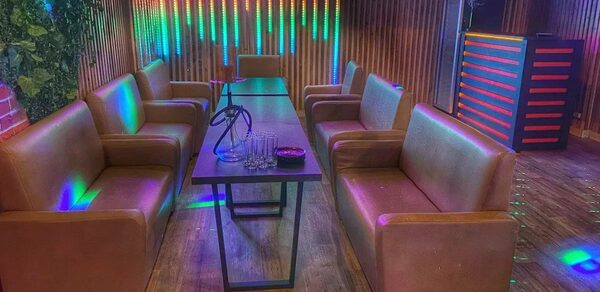 Forest(Лес). Простор с деревянными стенами и полом, длинный стол, окружённый коричневыми диванами, кальян, бокалы и пепельница на столе. На заднем фоне светодиодные панели с разноцветной подсветкой.