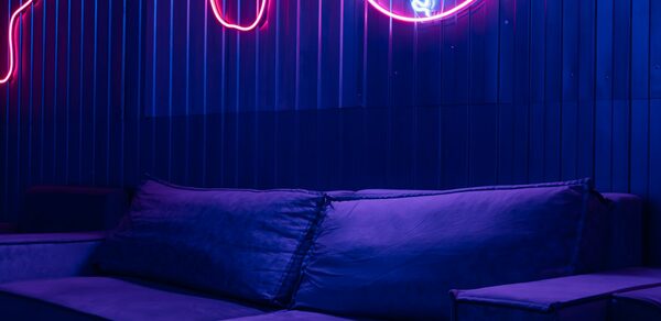 Rubin Loft (Рубин Лофт). Зал Токио. Лофт-пространство с темной мебелью, включая диван с подушками. Стена покрыта темными панелями, освещена неоновыми вывесками. Основные цвета — синий и сиреневый, создающие атмосферное освещение.