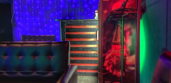 Rave(Рейв). Зона отдыха с черными кожаными диванами, барной стойкой с красными горизонтальными линиями, холодильником Coca-Cola, фоновым освещением в виде синих гирлянд, зелеными и красными световыми акцентами. Стены зелёного оттенка.