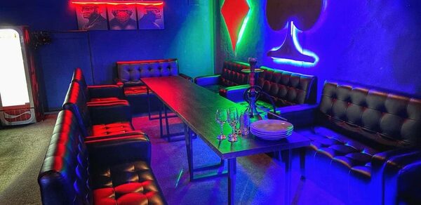 Rave(Рейв). Длинный стол в центре с черными кожаными креслами и диванами по периметру. Неоновое освещение: красный, зеленый, синий. На стене светящиеся символы карточных мастей. На столе набор бокалов и тарелок. Левый угол с холодильником.