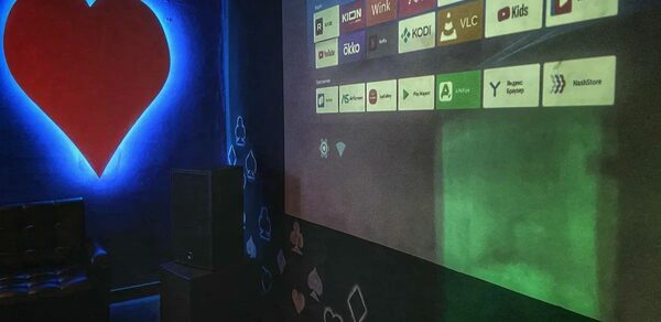 Rave(Рейв). Комната с проектором, на стене отображены иконки приложений. В левом углу большое красное сердце на синем фоне. Черная стена с белыми и красными фигурками в форме крестов и бубен. Справа темное кресло.