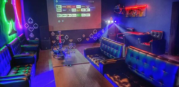 Rave(Рейв). Лофт-пространство с черными кожаными диванами, низким деревянным столом, проектором и экраном, столом для покера. На стенах неоновые знаки и картины. Освещение разноцветное, преимущественно синие и фиолетовые оттенки. Курительные принадлежности на столе.