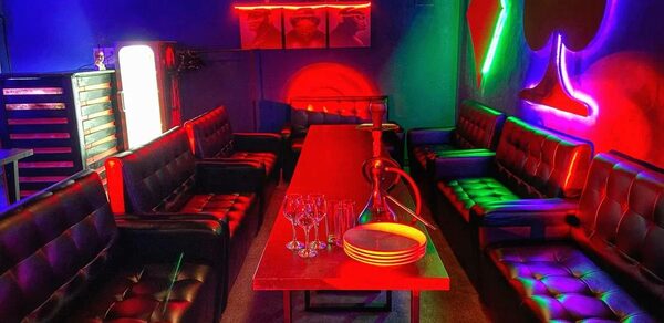 Rave(Рейв). Караоке-зал с длинным деревянным столом, окруженным кожаными диванами. На столе кальян и бокалы. Неоновые огни в виде мастей карт на стенах. Яркое разноцветное освещение, синий и красный интерьер. Декорировано постерами.