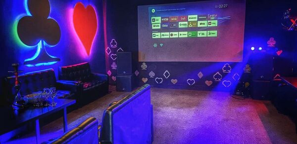 Rave(Рейв). Темное помещение с проектором на стене, показывающим экран интерфейса. Декор в виде светящихся знаков игральных карт. Мягкие диваны черного цвета, низкий стол с графинами и бокалами. Настенное освещение синих и красных оттенков.