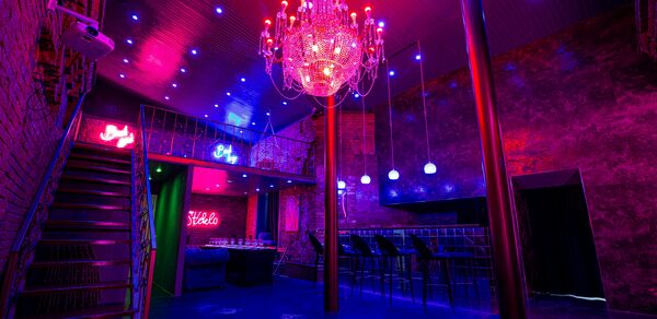 Двухэтажный лофт с отдельным входом. Двухуровневое помещение с кирпичными стенами и металлической лестницей. Потолок с точечным и неоновым освещением, люстра. Барная стойка с высокими стульями. На стенах неоновые надписи. Темно-красные и синие оттенки.