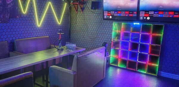 Bee-lounge(Пчела-лаундж). Караоке-зал с мягкими диванами и длинным деревянным столом. На стенах неоновая подсветка и светодиодные панели. Два телевизора на стене. Темный пол и сетчатые обои. Стеклянные стаканы и кальян на столе.