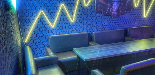 Bee-lounge(Пчела-лаундж). Кожаные диваны и прямоугольный стол в углу помещения. Синие стены с шестигранным узором. Светодиодные желтые неоновые линии на стене. Полумрак, освещение неоновое. Состояние мебели и отделки хорошее.