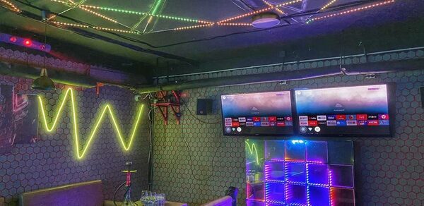 Bee-lounge(Пчела-лаундж). Караоке-зал с неоновыми огнями, светодиодными лентами на потолке, мягкими диванами и деревянными столами. На стене два телевизора, светящаяся панель и декоративные элементы. Стены декорированы узорчатой плиткой. На столе кальян и бутылки с водой.