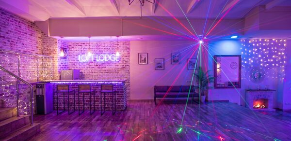 Loft Lodge (Лофт Лодж) 7. Просторный уютный лофт. Лофт-пространство с кирпичными стенами и деревянным полом, барная стойка с табуретами, неоновая надпись, световые эффекты, потолочные балки, декоративный камин, художественные рамки на стене, мягкая мебель, гирлянды из лампочек.
