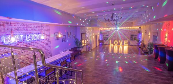 Loft Lodge (Лофт Лодж) 7. Просторный уютный лофт. Просторное помещение с кирпичными стенами и деревянным полом. Слева барная стойка и высокие стулья. Сверху подвесные светильники. В центре помещения диско-шар и мерцающие огни. На заднем плане большие световые буквы «LOVE».
