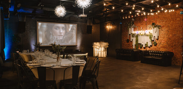 Nord Loft(Норд Лофт). Лофт-пространство с кирпичной стеной и индустриальным потолком. Стол с белой скатертью и стульями, кожаные диваны. Подвесные светильники и гирлянды. Проектор и экран. Неоновая вывеска и декоративные элементы на стене. Тёплый свет.