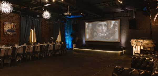 Nord Loft(Норд Лофт). Просторное праздничное помещение с кирпичными стенами и темным потолком. Длинный стол с белыми скатертями и стульями справа, кожаный диван слева. На стене большой экран с проектором. Подвесное освещение с гирляндами и декоративными светильниками.