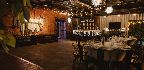 Nord Loft(Норд Лофт). Лофт-пространство с кирпичными стенами, круглым столом, черными стульями, кожаными диванами. Барная стойка с подсветкой, декор бутылками. Потолочные светильники в виде звезд. Стена с зеленью, светодиодные гирлянды, элементы неоновой вывески.