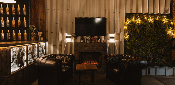 Nord Loft(Норд Лофт). Лофт-пространство с камином, два кожаных кресла, журнальный столик. На стене телевизор, рядом подсвеченные бутылки и живая стена с растениями. Освещение мягкое, с использованием подвесных ламп. Интерьер темный, с элементами из дерева и мрамора.