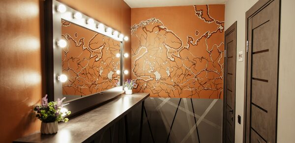 Nord Loft(Норд Лофт). Гримерная комната с большим зеркалом, обрамленным лампочками. На стене изображена карта. Стены и двери темного цвета. На длинной столешнице стоит вазон с цветами. Потолок светлый с несколькими встроенными светильниками.