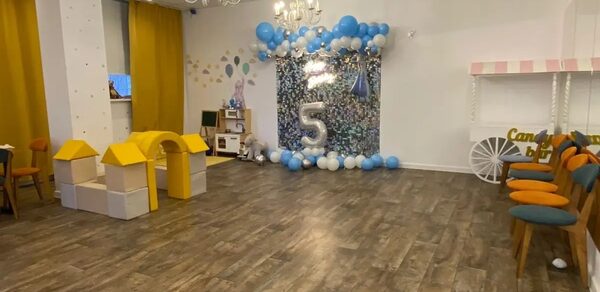 Лофт с потолками 3.5 м. просторное помещение с деревянным полом, белыми стенами и несколькими люстрами. У стены праздничная инсталляция с шарами и цифрой 5. Сбоку детские игрушечные домики и ряды стульев. Светлое оформление с жёлтыми и голубыми акцентами.