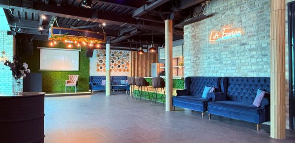 Лофт Фантом. Большое открытое помещение с кирпичными стенами и деревянным полом, декорированное неоновым светом. Синие диваны, барные стулья. На стенах зелёные панели и рисунки. Экран для проекций. Потолок с чёрными балками и светильниками.