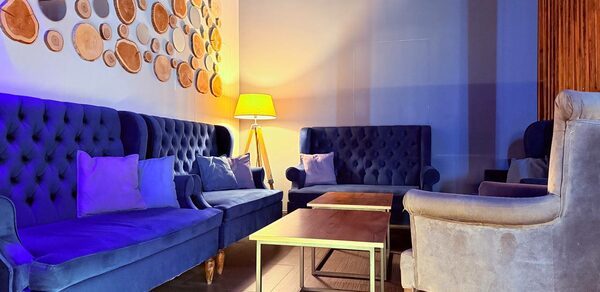 Лофт Фантом. Зал с тёмно-синими бархатными диванами и креслами, деревянными журнальными столиками, декоративной стеной с деревянными кружками. Напольная лампа с жёлтым абажуром создает мягкое освещение. Стены светлые, пол деревянный.