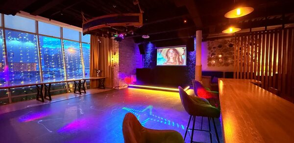 Лофт Фантом. Просторное помещение с барной стойкой, высокими стульями и зоной танцпола. На стене большой экран. Окна от пола до потолка украшены гирляндами. Потолок тёмный, полы гладкие, освещение разноцветное. Стены из кирпича и дерева.