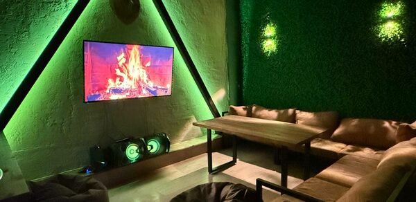 Лофт Фантом. Лаунж-зона с угловым диваном и деревянным столом, стены из штукатурки и зелени. На стене телевизор с изображением костра. На полу пуфики. Освещение зелёное, современная аудиосистема. Стены декоративные, разные фактуры.
