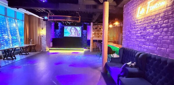Лофт Фантом. Клубное пространство с деревянным полом. Левая стена — окна с тёмными шторами. Справа кирпичная стена с неоновым знаком. У дальней стены сцена с экраном. Мягкие диваны и стулья, неоновая и точечная подсветка.