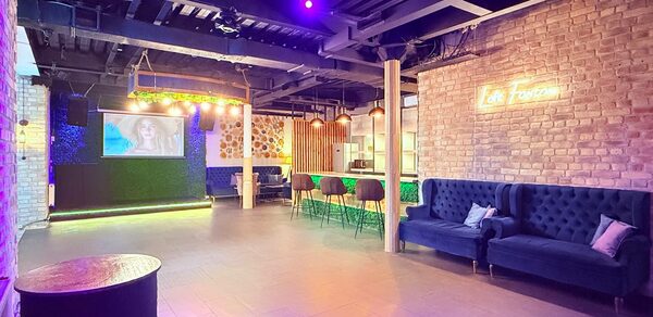 Лофт Фантом. Лофт-пространство с деревянными и кирпичными стенами; большая центральная зона с синими диванами; барная стойка с высокими стульями у стены; подсветка фиолетового оттенка; на стене надпись неоновыми буквами.