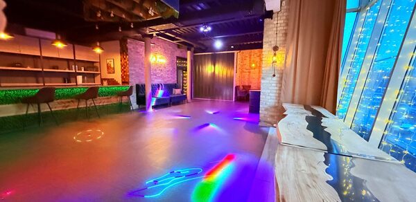 Лофт Фантом. Просторное помещение с танцполом и неоновым светом, барная стойка с высокими стульями, стол из светлого дерева, кирпичная стена, большие окна с гирляндами огней, черный потолок с балками, разноцветные световые узоры на полу.