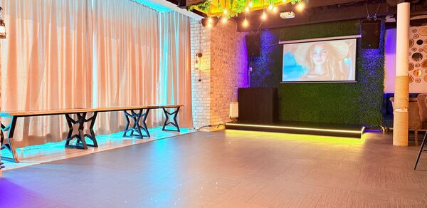 Лофт Фантом. Просторное помещение с деревянным паркетом и светодиодным освещением. Белые кирпичные стены, большие светлые шторы, деревянные столы. На одной стене экран с проектором, оформленная зелёной растительностью. Потолок декорирован светодиодными лампами.