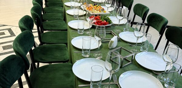 Arcobaleno Loft(Аркобалено Лофт). Длинный банкетный стол с зеленой скатертью и рядами темно-зеленых стульев, сервирован белыми тарелками, бокалами и приборами. На середине стола блюда с закусками. Пол с узором светлых и темных плиток.