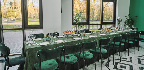 Arcobaleno Loft(Аркобалено Лофт). Пространство с длинным столом, покрытым зеленой скатертью, окружено стульями с зелеными бархатными сиденьями. На столе стеклянные бокалы, белые тарелки и закуски. Большие окна с видом на парк. Пол выложен узорчатой плиткой.