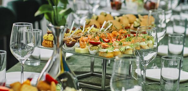 Arcobaleno Loft(Аркобалено Лофт). Длинный банкетный стол с зелёной скатертью, сервирован тарелками с разнообразными канапе, бокалами для вина и стеклянными стаканами. Вазы с цветами украшают стол. Цвета: зелёный, белый, металлические элементы.