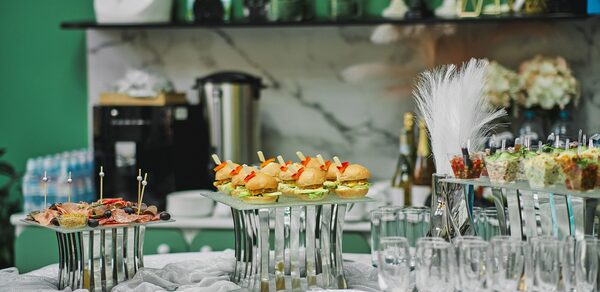 Arcobaleno Loft(Аркобалено Лофт). Праздничное пространство со столом для фуршета. На столе бутерброды, закуски в стеклянных стаканах, десерты, стеклянные бокалы. На заднем плане декор и напитки, зеленые стены, бело-мраморная столешница.