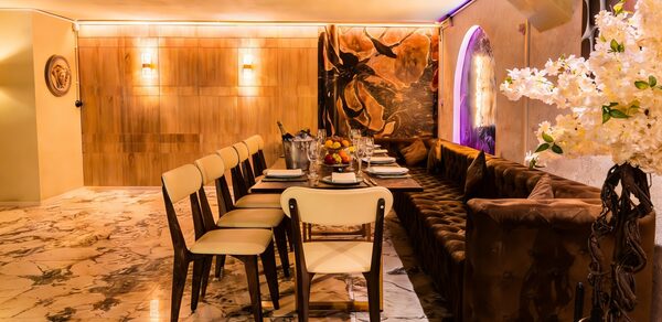 Стильный лофт Джаванди. Современное помещение с мраморным полом. Длинный стол с сервировкой и восемью стульями с бежевыми сиденьями. Справа диван, в углу ваза с белыми цветами. На стене деревянные панели и декоративная картина. Мягкое освещение.