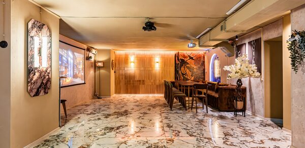 Стильный лофт Джаванди. Просторное помещение с мраморным полом, деревянной стеной, несколькими столами и стульями. На стенах висят картины и светильники. Потолочный проектор. Декоративные цветы и растения. Тёплое освещение.