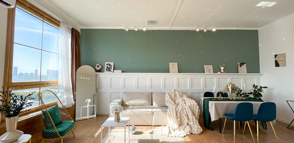 Зал Квартира. Уютное пространство в квартирном стиле. Гостиная с деревянным полом, зелёной акцентной стеной и белыми панелями. Диван с пледом, кофейный столик, обеденный стол с синими стульями, зеркало, растения. Большое окно с видом на город. Естественное и трековое освещение.