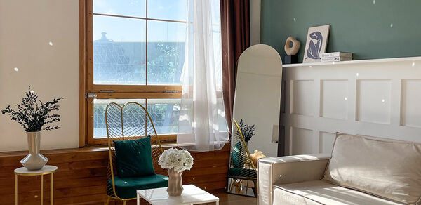 Зал Квартира. Уютное пространство в квартирном стиле. Светлая комната с большим окном, белыми занавесками и деревянными элементами. Белый диван, два небольших столика с цветами, декоративное зеркало, зелёное кресло с подушкой. Светло-зелёная стена, частичная деревянная панель.