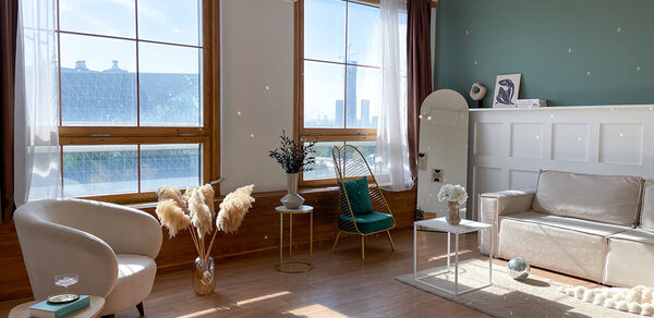 Зал Квартира. Уютное пространство в квартирном стиле. Светлая комната с большими окнами, деревянным полом и белыми стенами. Мебель: диван, два кресла, декоративные столы. Украшения: вазы с сухими травами, зеркало. Освещение естественное. Кондиционер на стене, зеленая акцентная стена.