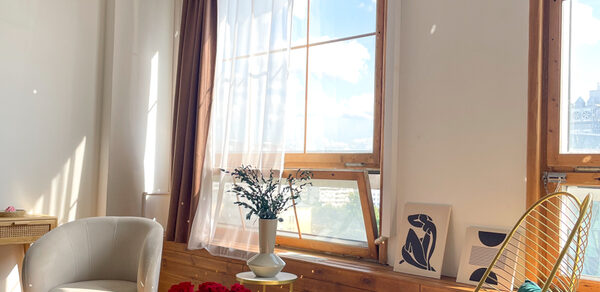 Зал Квартира. Уютное пространство в квартирном стиле. Уютная комната с большими окнами и прозрачными занавесками, деревянными подоконниками. Белое кресло, стеклянный стол с красными розами и ваза с цветами. Светлые стены, деревянные элементы, картина на подоконнике, солнечное освещение.