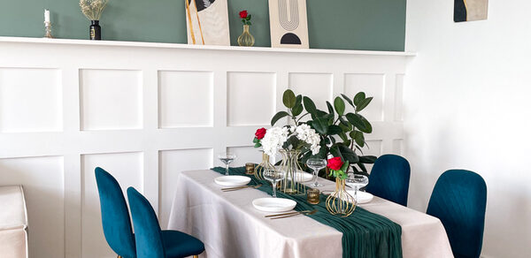 Зал Квартира. Уютное пространство в квартирном стиле. Столовая зона с белым панельным оформлением стен, обеденным столом с белой скатертью, украшенной зелёным текстилем и цветочными композициями. Синие стулья, деревянный пол, картины и декор на стене. Светлая и уютная атмосфера.