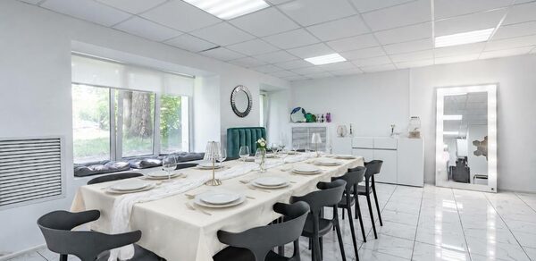 Handy Class (Хэнди Класс). Современный зал. Современное светлое помещение с длинным столом, накрытым белой скатертью, и черными стульями. Окно с видом на природу, белые стены и плиточный пол. Зеркало на стене, настольные лампы, декор в виде растений, потолочные светильники.