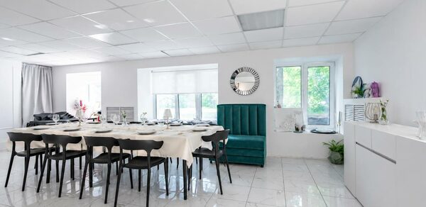 Handy Class (Хэнди Класс). Современный зал. Белая комната с плиточным полом и подвесным потолком. Длинный стол, покрытый белой скатертью, окружен черными стульями. Зеленый диван вдоль стены, зеркало и два окна. Белый шкаф, декоративные растения. Светлое естественное освещение.