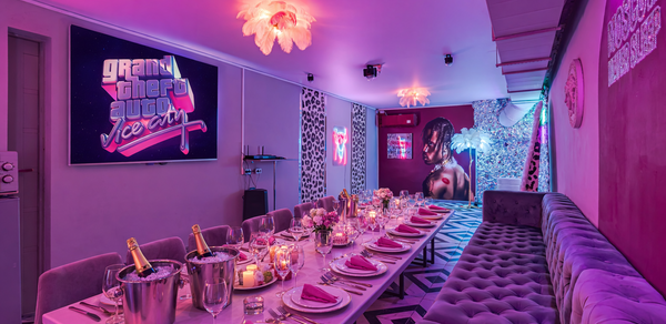 Неоновый лофт Гламур. Ресторан с длинным столом, сервированным бокалами и салфетками. Стены декорированы постерами и неоновыми вывесками. Мягкий диван вдоль стены. Потолочное освещение с лампами в виде цветов. Интерьер в розово-фиолетовых тонах.