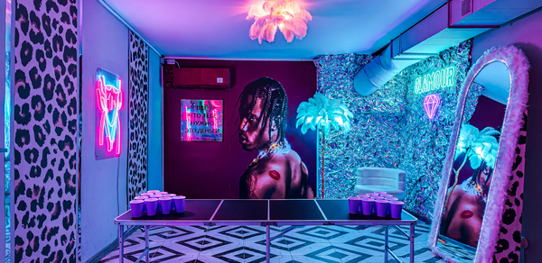 Неоновый лофт Гламур. Квадратная комната с шахматным полом, неоновыми вывесками, зеркалом в яркой раме и большим постером на стене. В центре стол с пластиковыми стаканами. Освещение розово-фиолетовое, на потолке светильник, похожий на перья.