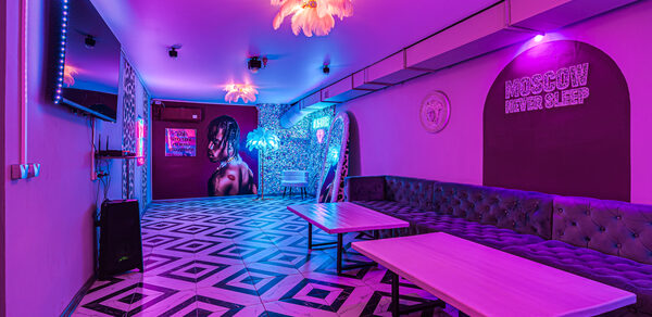 Неоновый лофт Гламур. Развлекательное пространство с геометрическим полом, фиолетовым освещением и неоновыми вывесками. Длинный серый диван вдоль стены, два прямоугольных стола. Декоративные светильники на потолке, настенные изображения и растения с подсветкой.