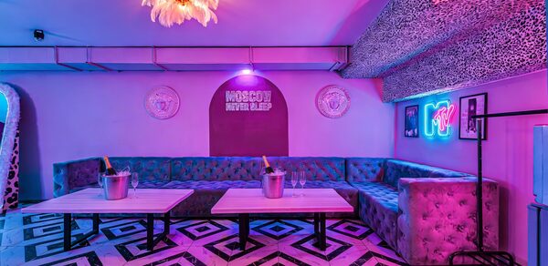 Неоновый лофт Гламур. Караоке-зал с угловыми диванами обития, два стола с ведёрками шампанского, плиточный пол с геометрическим рисунком, неоновая надпись, яркая люстра, фиолетово-розовая подсветка, леопардовый принт на потолке, круглые зеркала, декорированные стены.