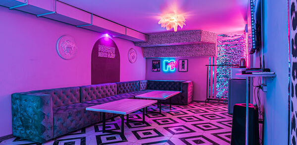 Неоновый лофт Гламур. Караоке-зал с неоновой подсветкой, фиолетовые и розовые световые акценты. Угловой диван из зелёного бархата, два прямоугольных стола, черно-белый узорчатый пол, стены с декоративными элементами и телевизор. Табличка с надписью и неоновый логотип.