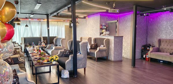 Зал с террасой до 55 гостей. Просторное помещение с серыми креслами, барной стойкой и столами. На стенах светлая текстура, потолок тёмный. Освещение светодиодное с фиолетовыми оттенками. Есть проектор, шары и сервированные столы с едой. Пол тёмного дерева.