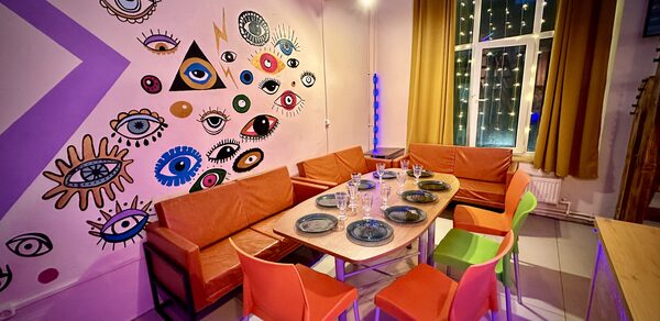 Яркий двухуровневый лофт. Стол с посудой. Яркие стулья с диванами. Обклеенные рисунками стена. Окно с навешенной гирляндой