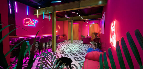 Плэйс Лофт. Prisma Loft(Призма Лофт). Барное пространство с розовыми стенами и неоновыми вывесками. Пол с черно-белой шахматной плиткой. Зона с барной стойкой и высокими стульями. Мягкая мебель, декоративные растения, яркое разноцветное освещение. Шторы тёплых оттенков.